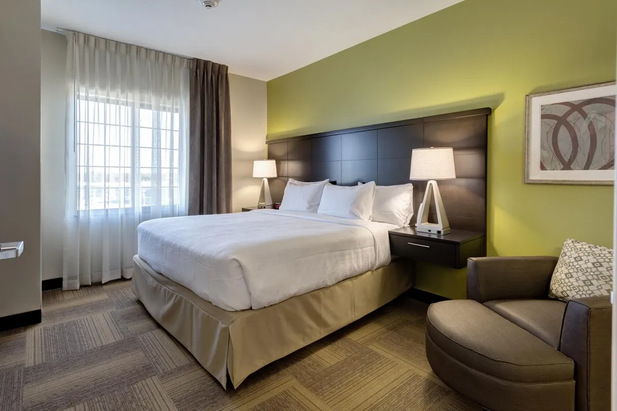 A serene bedroom featuring a plush king-size bed topped with crisp linens, flanked by modern nightstands and soft lamps. An accent chair sits beside a large window draped in sheer curtains, letting in diffused natural light. The restful color palette and uncluttered design create a sanctuary-like atmosphere, with subtle decor adding a touch of elegance.