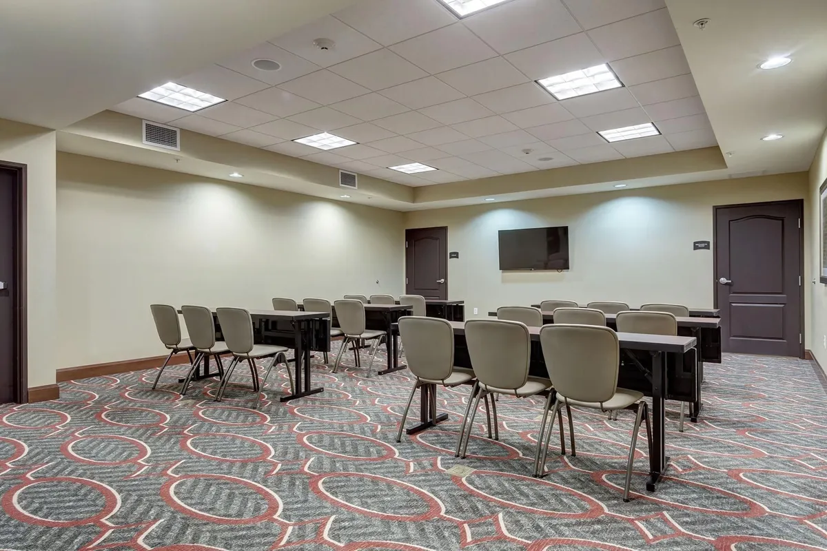 A spacious, well-lit interior setting featuring several tables and chairs arranged for group interactions. The room includes a modern television mounted on the wall and a distinctive carpet design, creating an inviting yet functional atmosphere. The furnishings and setup suggest it is suitable for meetings, collaborative activities, or social gatherings, reflecting a communal aspect of the property.