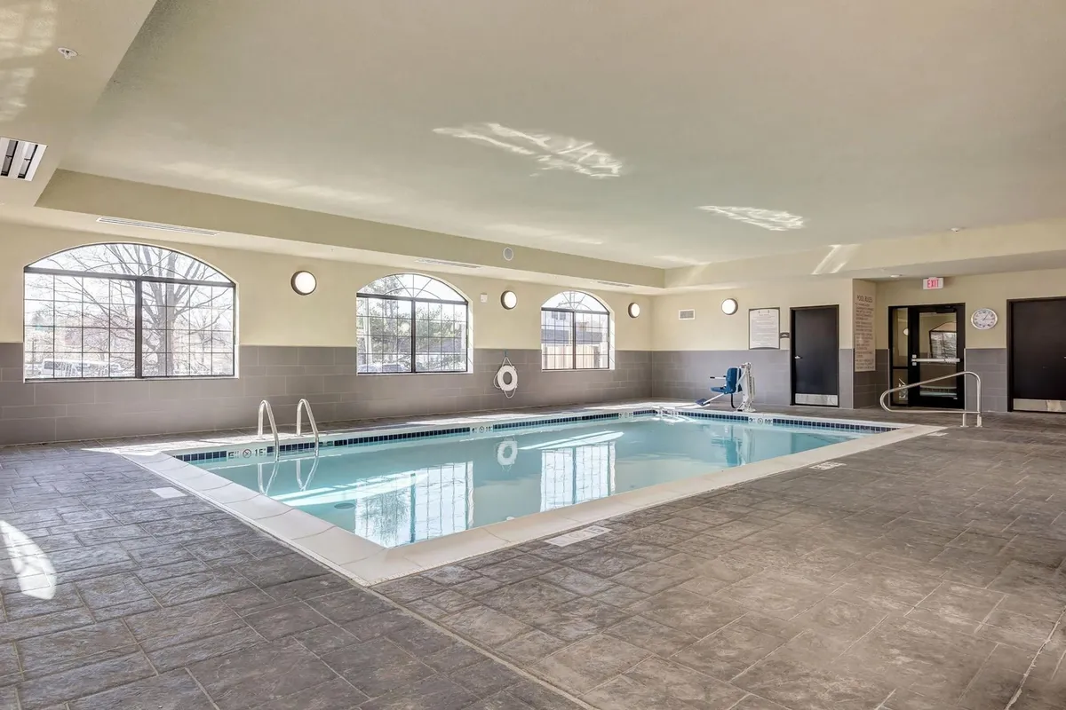 A spacious indoor swimming pool area filled with natural light from large windows. The pool is surrounded by comfortable poolside seating, and there are a few guests swimming and relaxing, evoking a calm and refreshing atmosphere.
